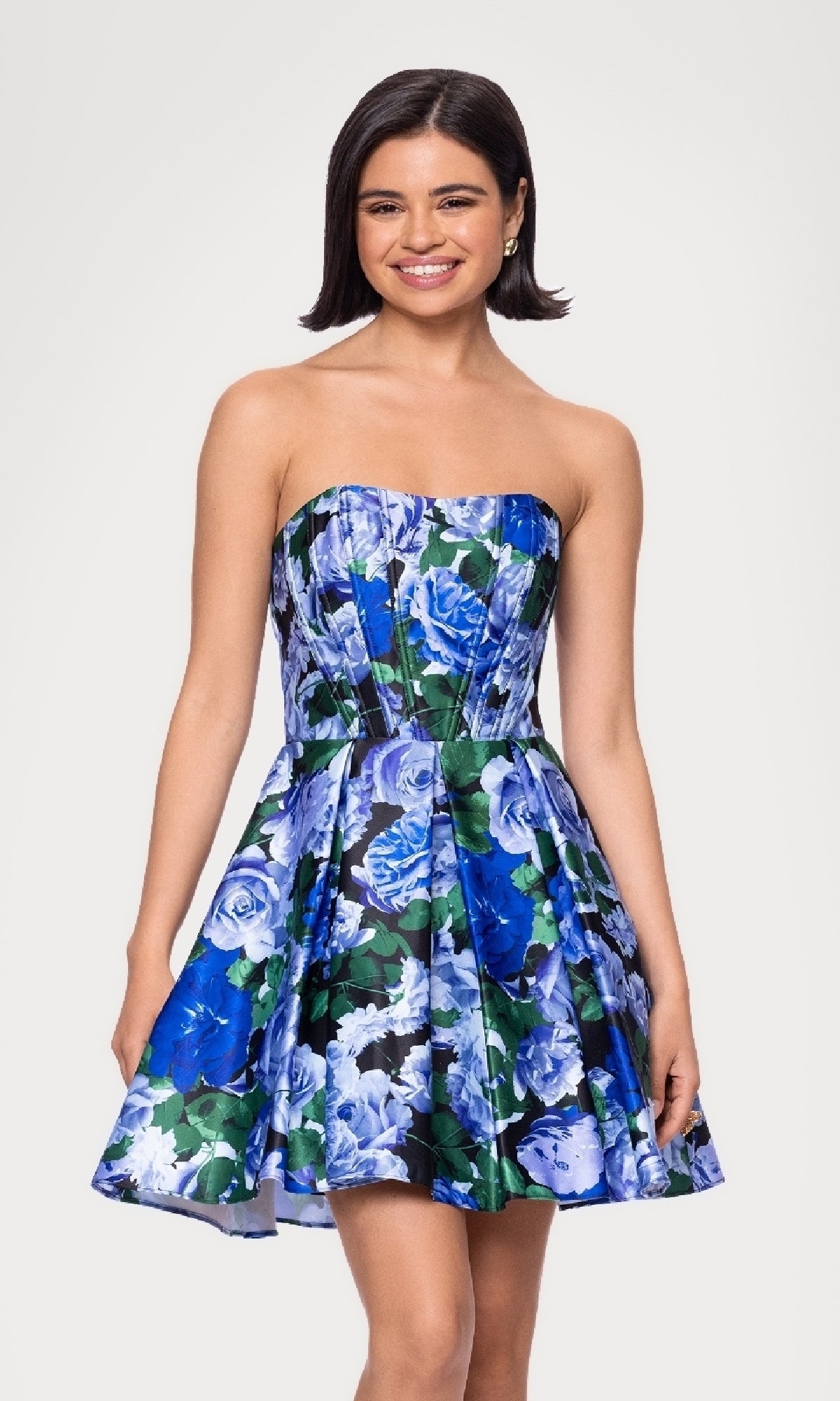 A woman wearing the Strapless Print Hoco Dress: Blondie Nites 5592BN, an affordable semi-formal knee-length strapless floral dress in blue and green, stands smiling against a plain white background.